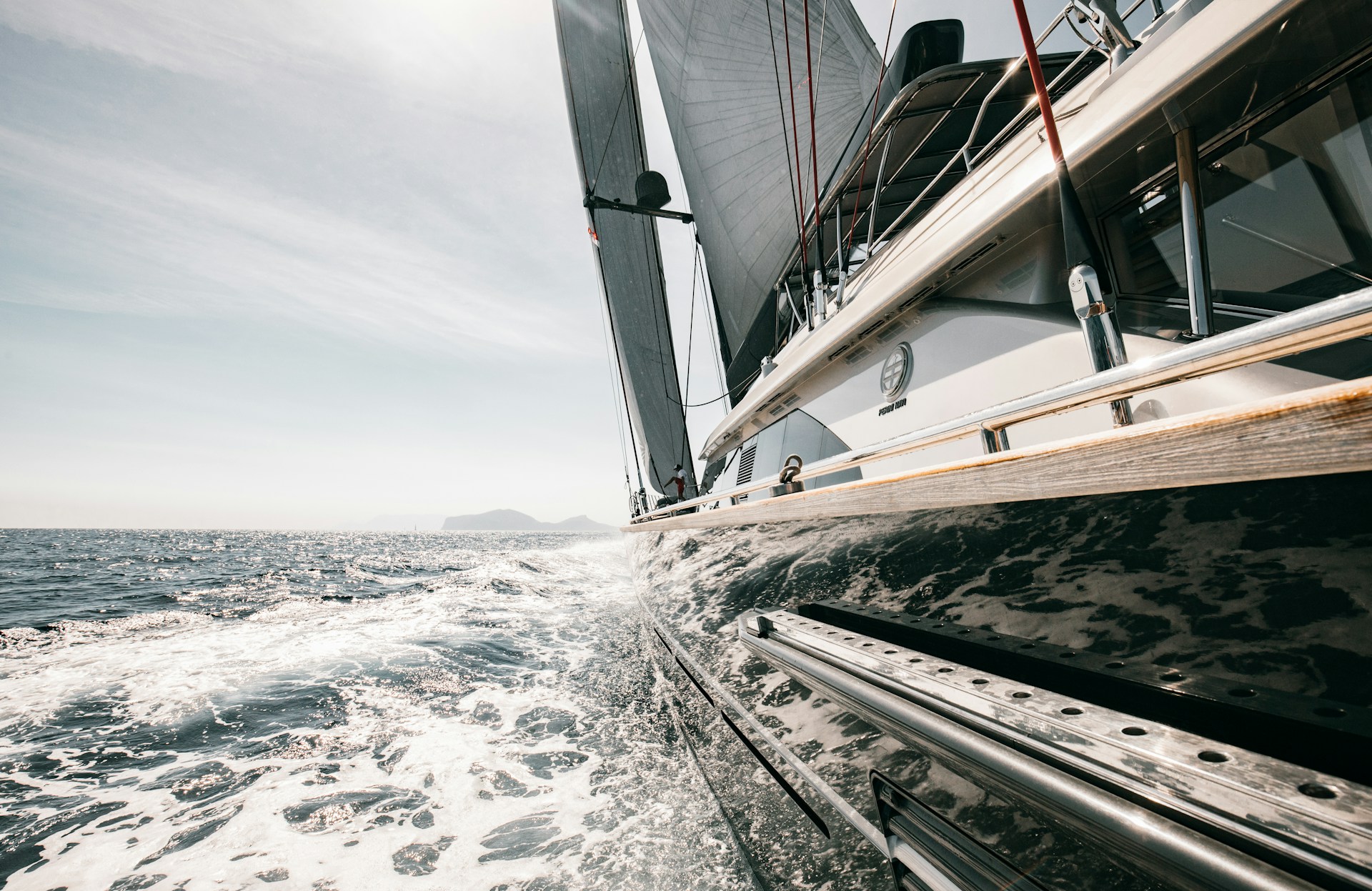 View from alongside a sailing yacht in motion, showing the vessel’s hull, rigging, and sails with seawater foaming alongside under a bright sky.