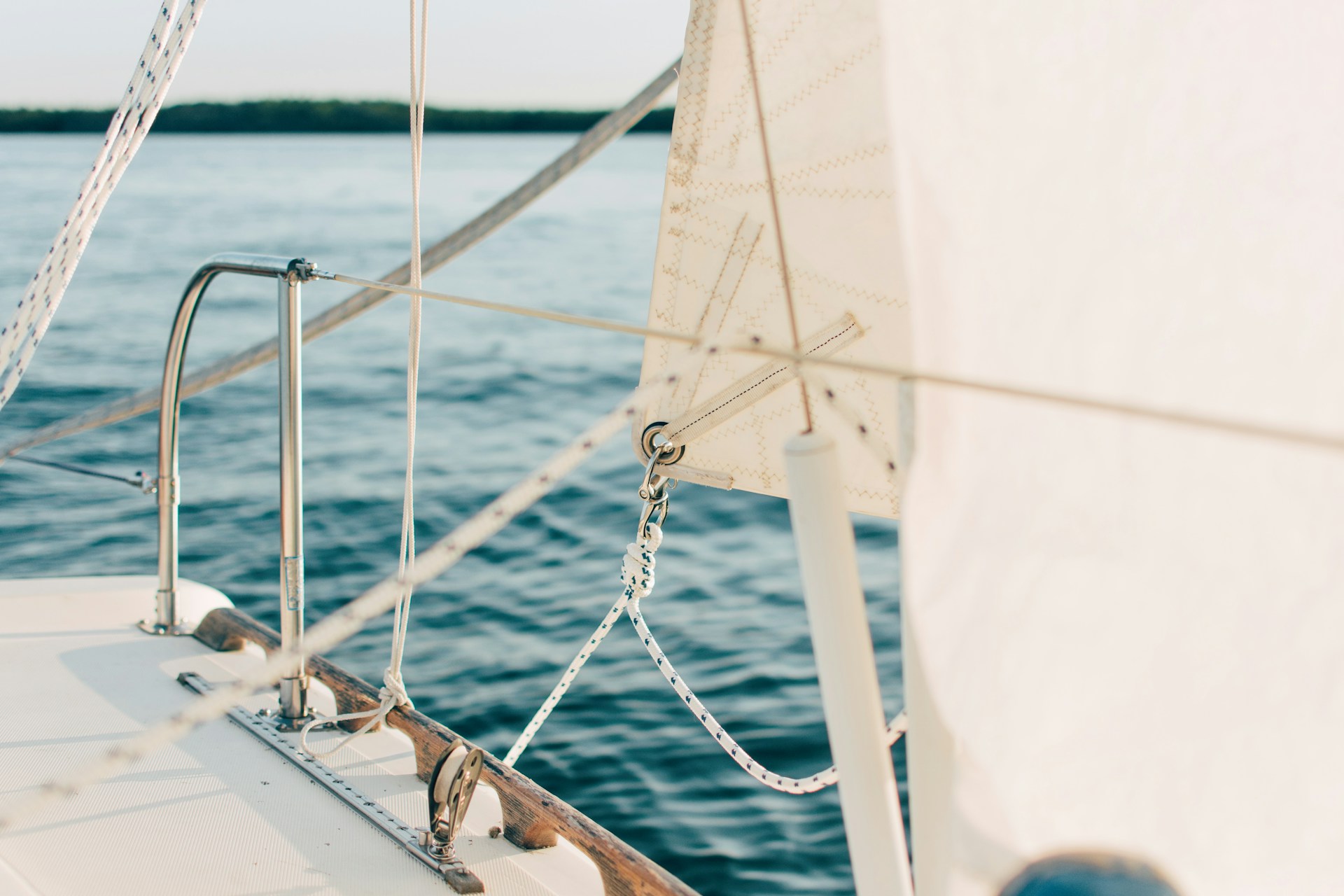 Close-up view of a sailboat deck with white sails, rigging lines, and guard rails visible beside calm blue water.