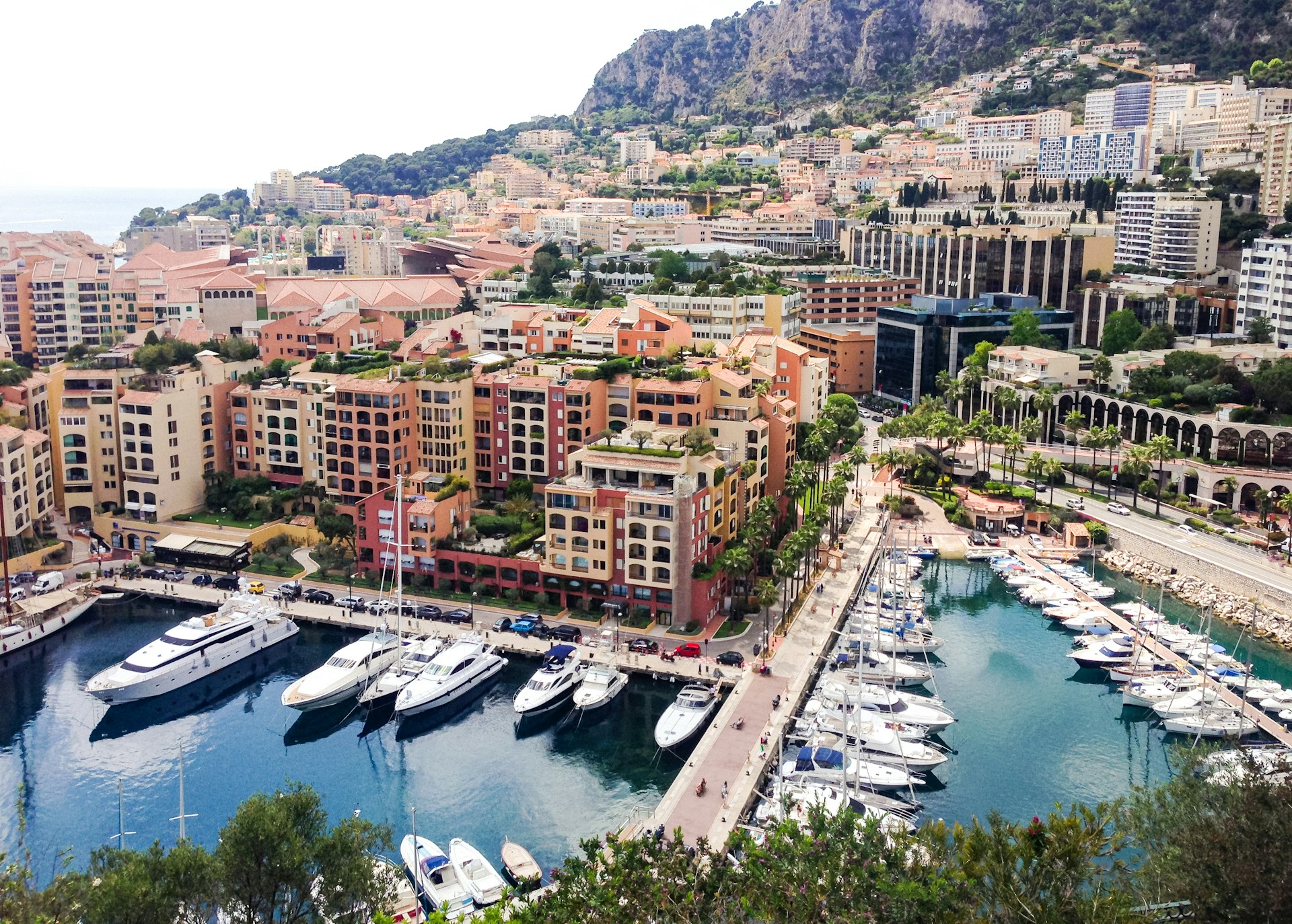 Aerial view of a waterfront marina filled with yachts and boats, surrounded by densely packed apartment buildings and streets, with steep hills rising in the background.